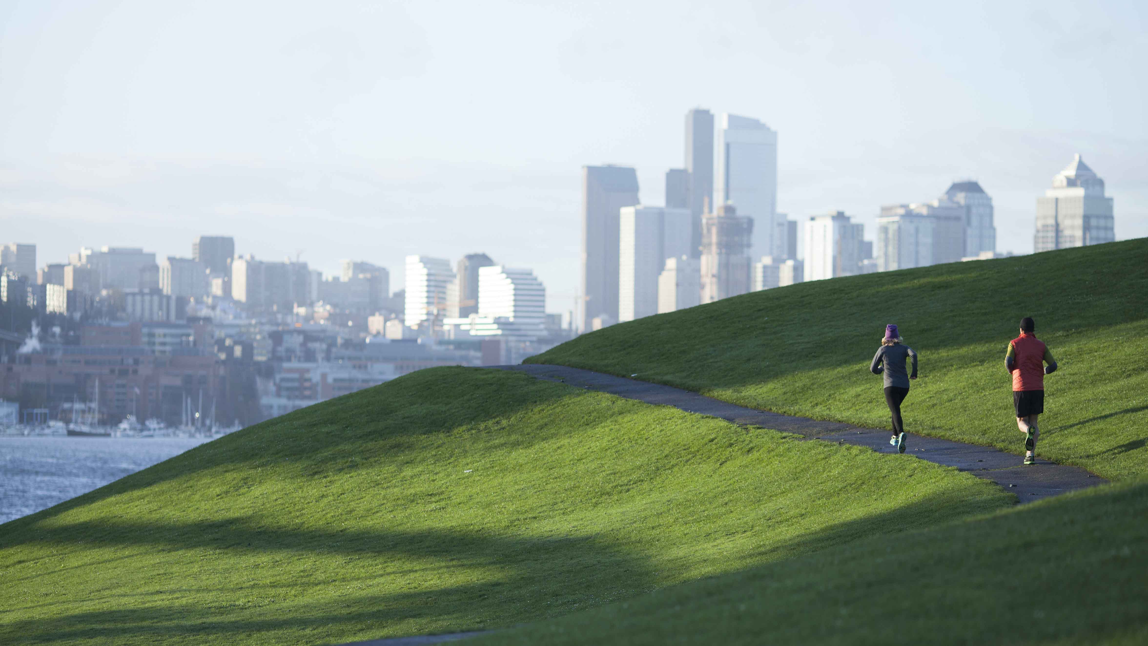 2 male and female joggers on a path through green field, overlooking city